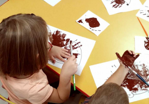 Kilkoro dzieci przy stoliku stempluje szablon jeża pomalowaną dłonią.