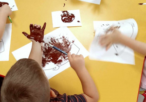 Kilkoro dzieci przy stole maluje dłonie pędzelkiem przy użyciu brązowej farby.