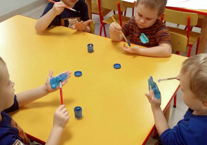 Kilkoro dzieci przy stoliku maluje dłonie pędzelkiem umoczonym w niebieskiej farbie.