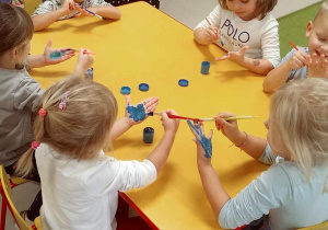 Kilkoro dzieci przy stoliku maluje dłonie pędzelkiem umoczonym w niebieskiej farbie.