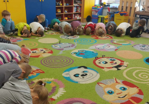 Dzieci z grup starszych siedzą na dywanie z zamkniętymi oczami podczas wypowiadania magicznego hasała zmniejszającego i zwiększającego misia Teddy Eddie.