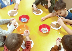 Kilkoro dzieci przy stoliku podczas ozdabiania pierników przy pomocy kolorowego lukru oraz posypek.