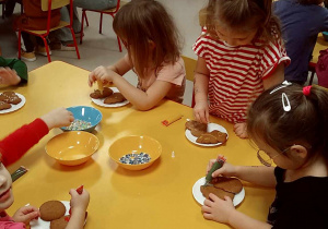 Kilkoro dzieci przy stoliku podczas ozdabiania pierników przy pomocy kolorowego lukru oraz posypek.