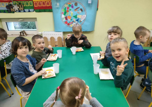 Kilkoro dzieci przy stole podczas zdrowego, drugiego śniadania. W tle jest konkursowa praca plastyczna wszystkich dzieci z naszej grupy - "Talerz zdrowia".