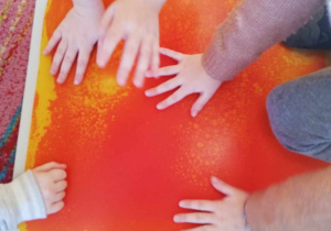 Kilkoro dzieci z grupy "Skrzatów" dotyka rączkami pomarańczowej płytki sensorycznej.
