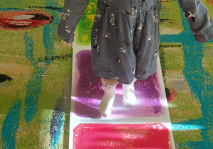 Dziewczynka z grupy "Biedronek" spaceruje po kolorowych płytkach sensorycznych na boso.