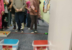 Zabawa ruchowa - "Rzuć kolor" - Klaudia ustawiona przed odpowiednim pojemnikiem rzuca piłką w kolorze pasującym do pojemnika. Pozostałe dzieci czekają na swoją kolej.