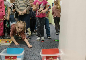 Zabawa ruchowa - "Rzuć kolor" - Roksana ustawiona przed odpowiednim pojemnikiem rzuca piłką w kolorze pasującym do pojemnika. Pozostałe dzieci czekają na swoją kolej.