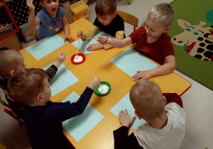 Kilkoro dzieci przy stole stempluje kartkę, wykorzystując poprzecinaną słomkę, umoczoną w białej farbie.