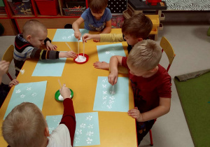 Kilkoro dzieci przy stole stempluje kartkę, wykorzystując poprzecinaną słomkę, umoczoną w białej farbie.