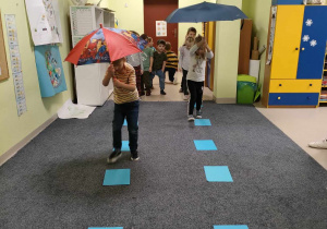 Artur i Klaudia podczas "deszczowego spaceru" z parasolami w dłoniach przeskakują przez niebieskie kartki(kałuże).