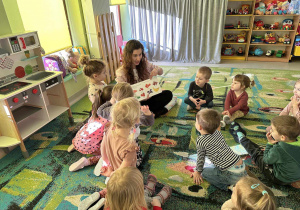 Pani Lidia pokazuje dzieciom ilustracje warzyw i owoców w książce. Dzieci siedzą na dywanie i uważnie oglądają.