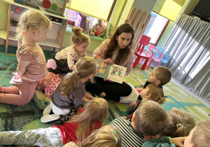 Mama Jagódki czyta dzieciom książeczkę pt. "Kicia Kocia. Witaminowe przyjęcie". Dzieci siedzą na dywanie i z uwagą słuchają.