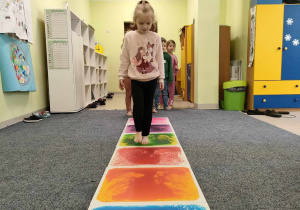Oliwia idzie stopa na stopą po kolorowej ścieżce sensorycznej.
