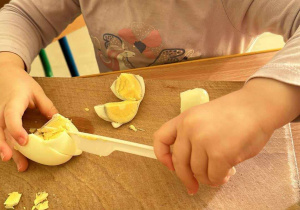 Przygotowanie sałatki brokułowej - Dziewczynka z maluszków kroi jajko na małe kawałeczki za pomocą plastikowego noża.