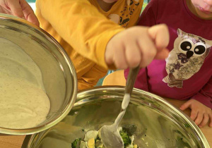 Przygotowanie sałatki brokułowej - Marika za pomocą łyżki dodaje do sałatki jogurt naturalny.