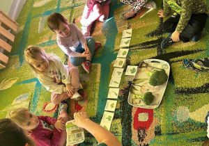 Ciocia Iwonka pokazuje dzieciom kiełki z brokuła. Dzieci siedzą w kole na dywanie.