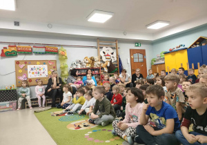 Przedszkolaki siedzą na dywanie i z uwagą oglądają występ dziewczynek.