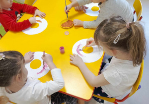 Kilkoro dzieci przy stole maluje wycięty szablon donata, przy pomocy kolorowej farby i pędzelka.