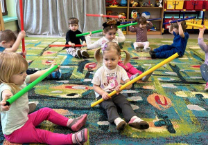Dzieci z grupy "Żabek" siedzą na dywanie i ćwiczą przy użyciu kolorowych lasek gimnastycznych.