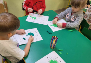 Kilkoro dzieci przy stole podczas pracy plastycznej, opartej na wyklejaniu kolorową plasteliną szablonu glutka.