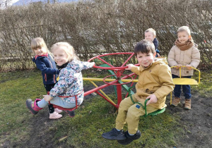 "Czas na aktywność ruchową na świeżym powietrzu" - kilkoro dzieci bawi się na karuzeli na przedszkolnym placu zabaw.
