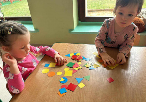 Jagódka i Amelcia przy stoliczku w trakcie matematycznych zabaw z kolorowymi figurami geometrycznymi.