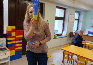 Pani logopeda - Magdalena Zdziarska prezentuje dzieciom ćwiczenie oddechowe z wykorzystaniem plastikowego kubka, gumowej rękawiczki i słomki.