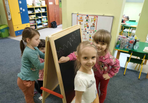 Marcelinka i Karolinka rysują kolorowymi kredami na czarnej tablicy.