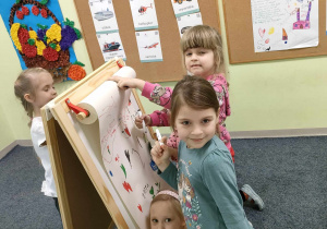 Karolinka, Zuzia i Marysia rysują kwiatki i serduszka kolorowymi mazakami na dużym arkuszu białego papieru.