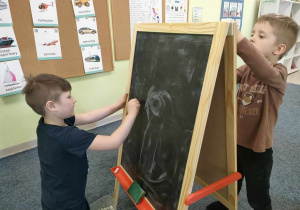 Alanek rysuje serduszko kolorową kredą na czarnej tablicy a Antoś tworzy swój rysunek kolorowym mazakiem na białym arkuszu papieru.