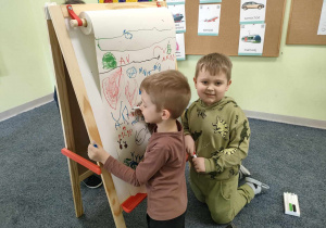 Marcelek i Antoś rysują kolorowymi mazakami na dużym, białym arkuszu papieru.