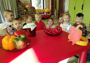 Paprykowy kącik wśród dzieci z grupy "Żabek" a w nim: papryki w trzech kolorach, papryczka chili, papryka konserwowa w słoiku oraz opaska z ilustracją czerwonej papryki.
