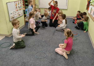 Ciocia Justynka tłumaczy dzieciom zasady zabawy orientacyjno-porządkowej "Tańczące jajka". Dzieci, które mają taki wzór pisanki, dobierają się w grupę.