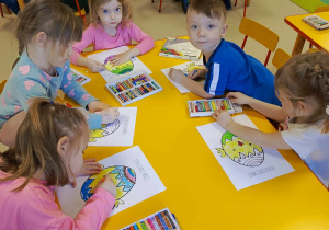 Kilkoro dzieci siedzi przy stoliczku i wypełnia szablon kurczaczka w pisance, przy wykorzystaniu kolorowych pasteli.