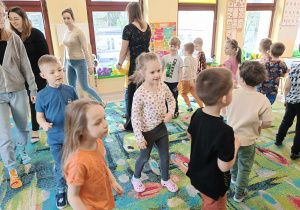 Dzieci wraz z zaproszonymi gośćmi chodzą po dywanie podczas zabawy ruchowej.