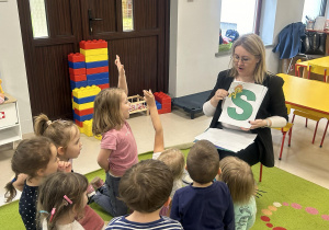Pani Ola pokazuje dzieciom z grupy młodszej szablon zielonej litery "S" z narysowaną pszczółką.