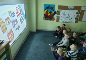Dzieci z grupy "Pszczółek" oglądają film edukacyjny pt. "Skąd się biorą produkty ekologiczne?" na tablicy interaktywnej.