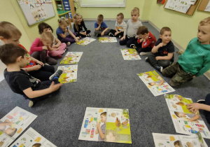 Dzieci z grupy "Pszczółek" siedzą w kole na dywanie i wspólnie oglądają publikację pt. "Skąd się biorą produkty ekologiczne?".