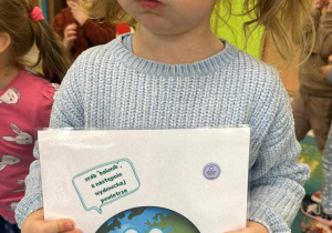 Dziewczynka trzymając kartkę z obrazkiem ziemi wykonuje podobną minę co planeta.