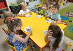 Kilkoro dzieci z grupy "Motylków" przy stoliku, podczas degustacji pysznej, słodkiej sałatki owocowej z dodatkiem soku z cytryny i miodu akacjowego.