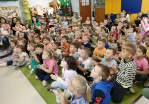 Dzieci siedzą na dywanie i słuchają śpiewu p. Emilki.