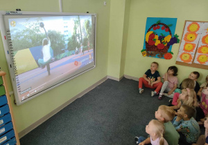 Dzieci starsze siedzą na dywanie i uważnie oglądają film edukacyjny o bezpiecznych wakacjach na tablicy interaktywnej.