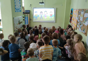 Dzieci starsze siedzą na dywanie i uważnie oglądają film edukacyjny o bezpiecznych wakacjach na tablicy interaktywnej.