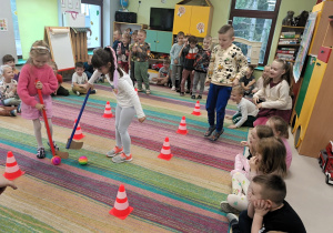 Dwie dziewczynki prowadzą piłkę za pomocą kija hokejowego między pachołkami. W tle widać chłopca, który mocno dopinguje swoją koleżankę z grupy.