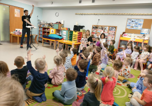 Dzieci siedzą na dywanie i aktywnie uczestniczą w muzycznym spotkaniu.