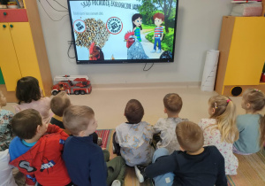 "Pszczółki" oglądają film animowany pt. "Skąd się biorą produkty ekologiczne?" na ekranie multimedialnym.