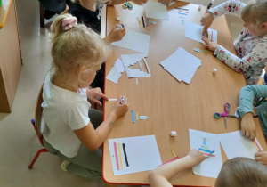Kilkoro dzieci przy stole nakleja elementy wg instrukcji słownej nauczycielki.