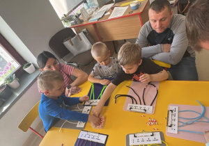 Kilkoro dzieci przy stoliczku wraz ze swoimi Opiekunami ćwiczy przeliczanie w zakresie 5-ciu, korzystając z kolorowych drucików kreatywnych.