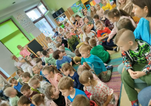 Przedszkolaki siedzą na dywanie i oglądają swoich rówieśników, którzy grają na instrumentach.